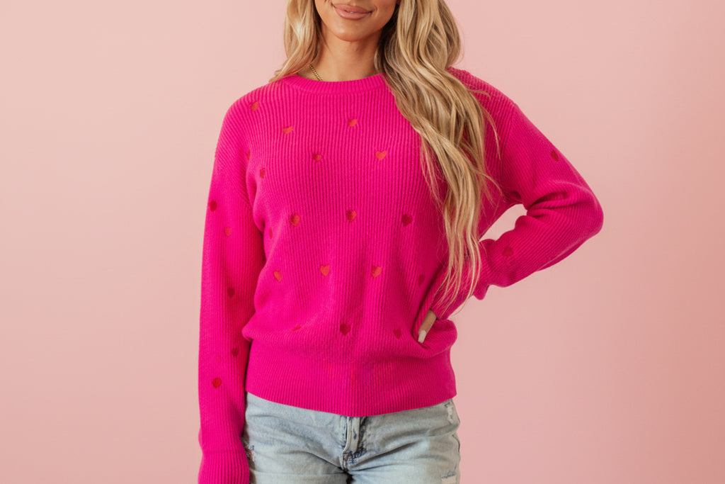 Person wearing a bright pink sweater with heart patterns, standing against a light background.